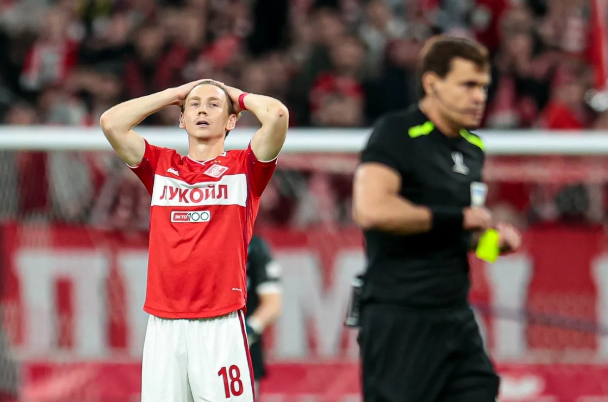 Момент футбольного матча — игрок в красном разочарованно реагирует на упущенный шанс, судья на заднем плане